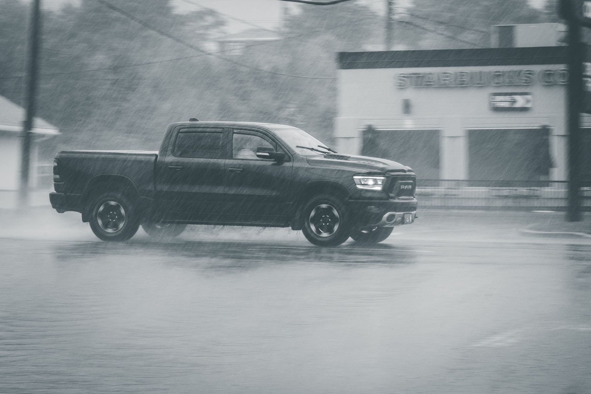 Ciemny pickup jedzie mokrą drogą w deszczu obok drzew i zabudowań